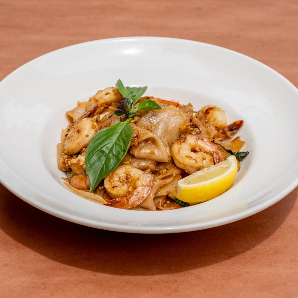 Pad See Ew with shrimp, basil, and a lemon wedge in a white bowl on a brown surface.