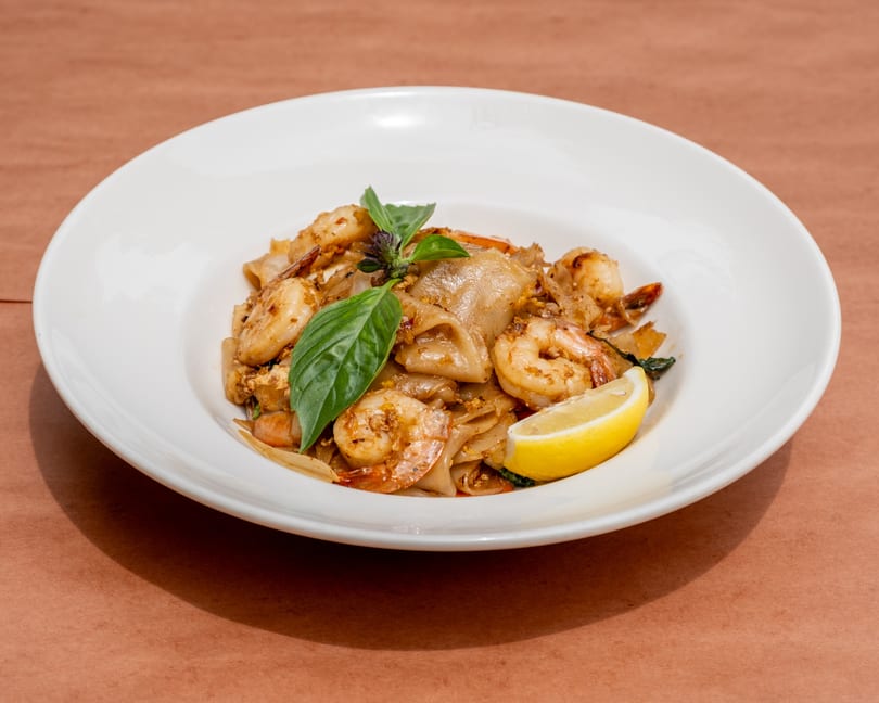 Pad See Ew with shrimp, basil, and a lemon wedge in a white bowl on a brown surface.