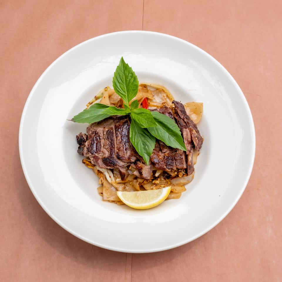 Sliced grilled steak with basil, wide noodles, and a lemon wedge on a white plate.