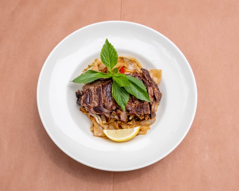 Sliced grilled steak with basil, wide noodles, and a lemon wedge on a white plate.