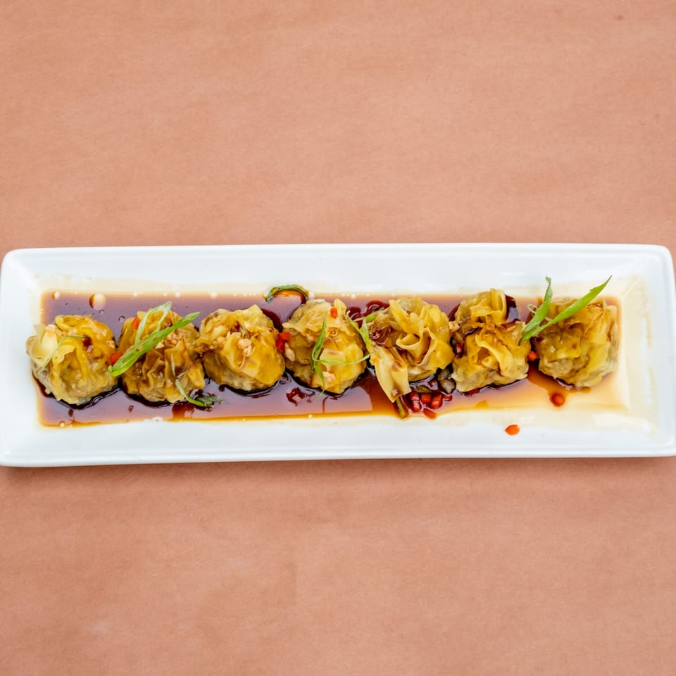 Seven dumplings in a dark sauce, garnished with green onions and red peppers, on a white rectangular plate.