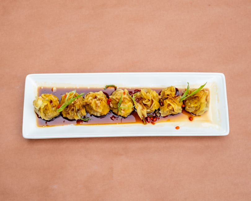 Seven dumplings in a dark sauce, garnished with green onions and red peppers, on a white rectangular plate.