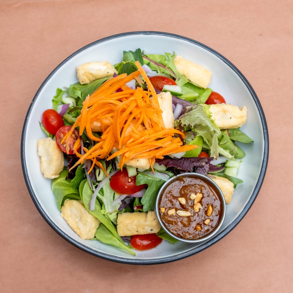 A vibrant salad with mixed greens, cherry tomatoes, cucumbers, shredded carrots, fried tofu, and peanut dressing.