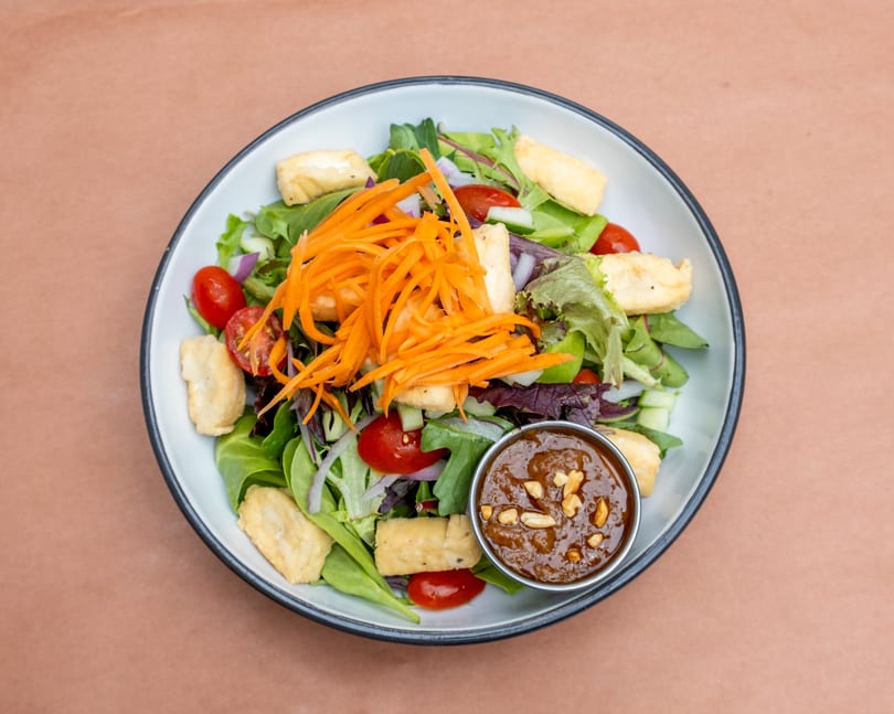 A vibrant salad with mixed greens, cherry tomatoes, cucumbers, shredded carrots, fried tofu, and peanut dressing.