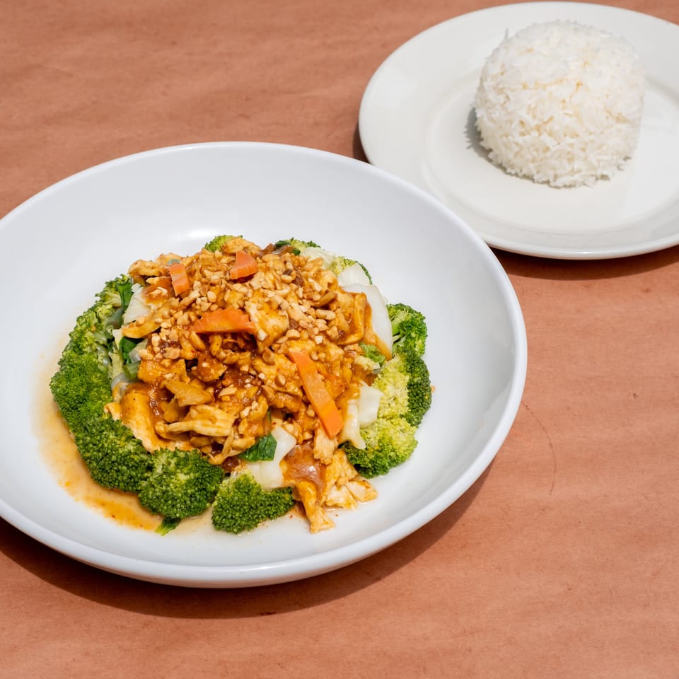 Chicken with peanut sauce, broccoli, and carrots in a white bowl, with a side of white rice.