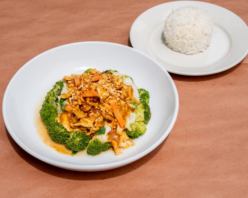 Chicken with peanut sauce, broccoli, and carrots in a white bowl, with a side of white rice.