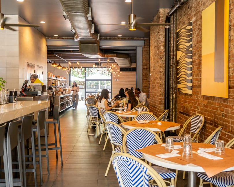 Restaurant interior with patrons dining at tables, a bar with stools, and exposed brick walls.