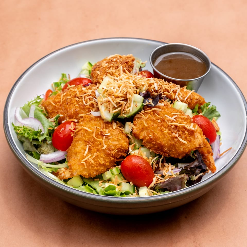 Coconut shrimp salad with mixed greens, cherry tomatoes, cucumbers, red onions, and a side of dressing.