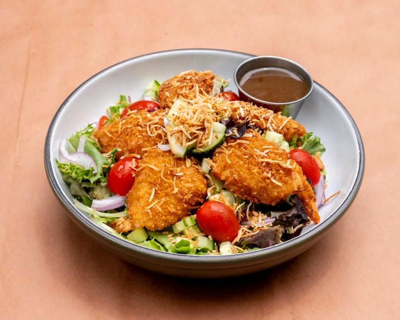 Coconut shrimp salad with mixed greens, cherry tomatoes, cucumbers, red onions, and a side of dressing.