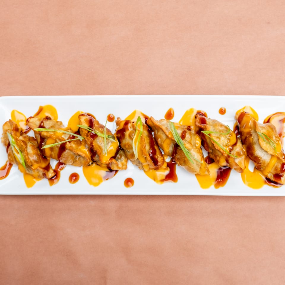 Fried dumplings drizzled with orange and brown sauces, garnished with green onions on a white plate.